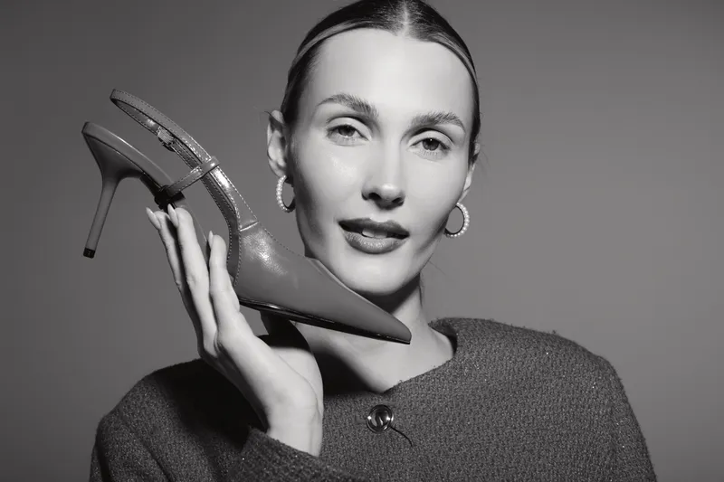 Young Caucasian woman with light skin holds high heel shoe near face, wearing hoop earrings and cozy sweater. Studio portrait in black and white, fashion and style concept.