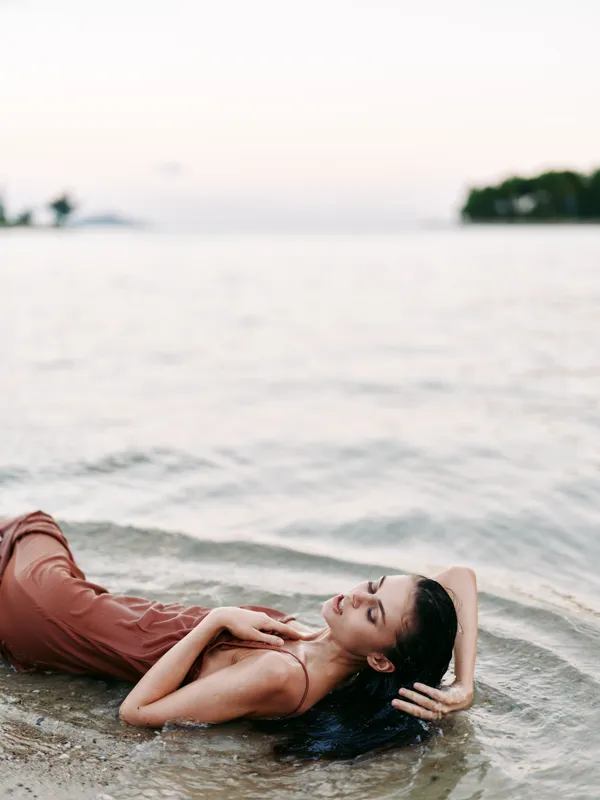 A woman lies in shallow sea water, relaxing in a rust dress as soft light brushes the beach. The tranquil scene focuses on calm body lines and serene shoreline ambiance.