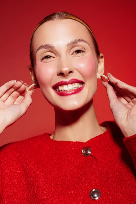 Smiling woman with shiny skin and red lipstick adjusts earrings on red background. She wears a red sweater with buttons. Concept of beauty, fashion, makeup, and joyful expression.