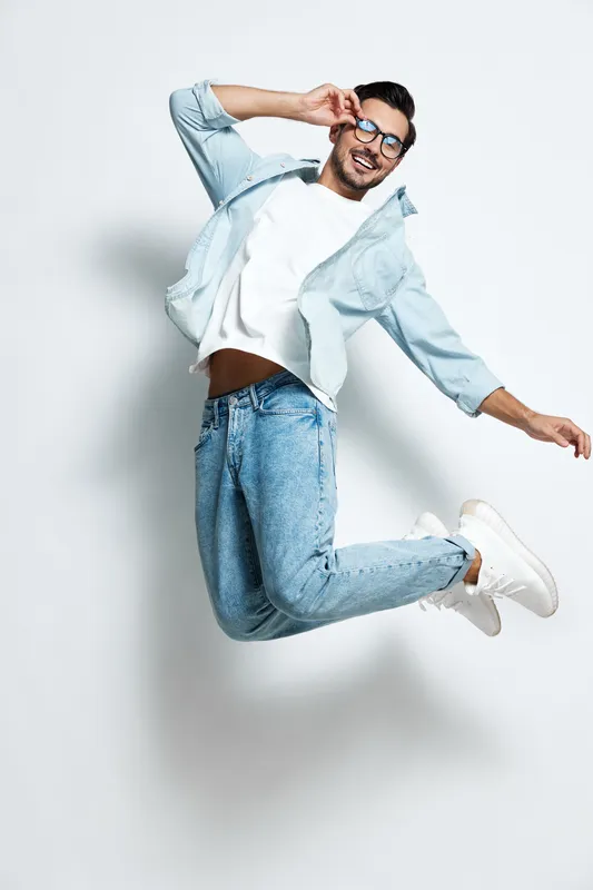 Full body joyful happy young man wearing denim jacket jumping in the air with a playful expression, wearing glasses, isolated on plain light background. Lifestyle concept.