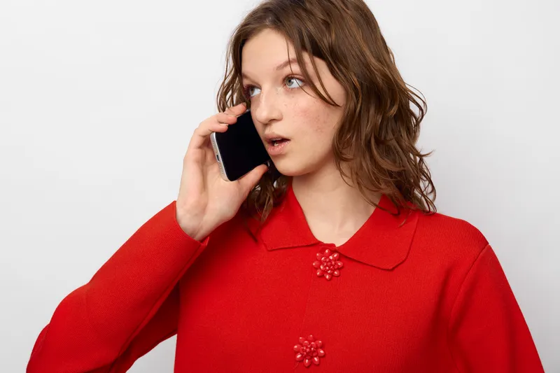 Young woman in red sweater talks on smartphone with surprised expression, isolated on white background. Concept of communication, technology, and unexpected news.
