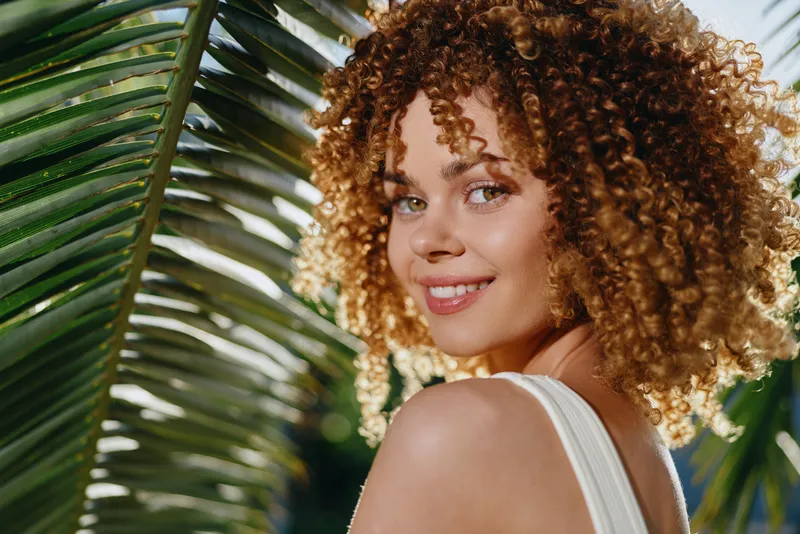 Smiling woman with curly hair poses outdoors near green palm leaves in natural sunlight, showcasing joyful expression and warm summer atmosphere.