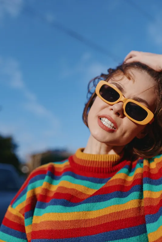 Woman smiling in a colorful striped sweater and yellow sunglasses, showing braces while tilting her head and touching hair outdoors in a sunny urban street for playful lifestyle fashion.