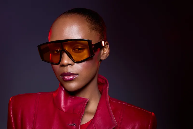 Portrait of confident African woman with dark skin wearing large orange sunglasses and red leather jacket. Studio shot with dramatic lighting and fashion concept emphasizing style and attitude.