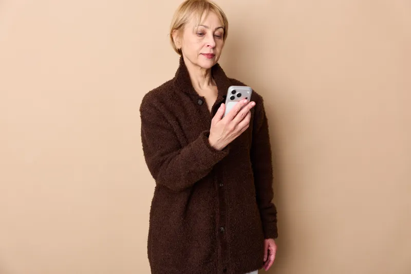 Elderly woman, smartphone, indoors, portrait, beige wall, winter coat, looking at screen, calm expression, soft studio light, hands holding device, copy space