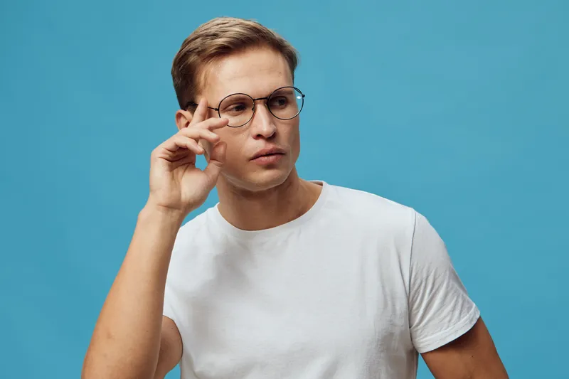 Thoughtful young Caucasian man with glasses wears white t-shirt against blue background, looks away, and adjusts glasses using hand, portraying concentration or reflection in a studio portrait. People