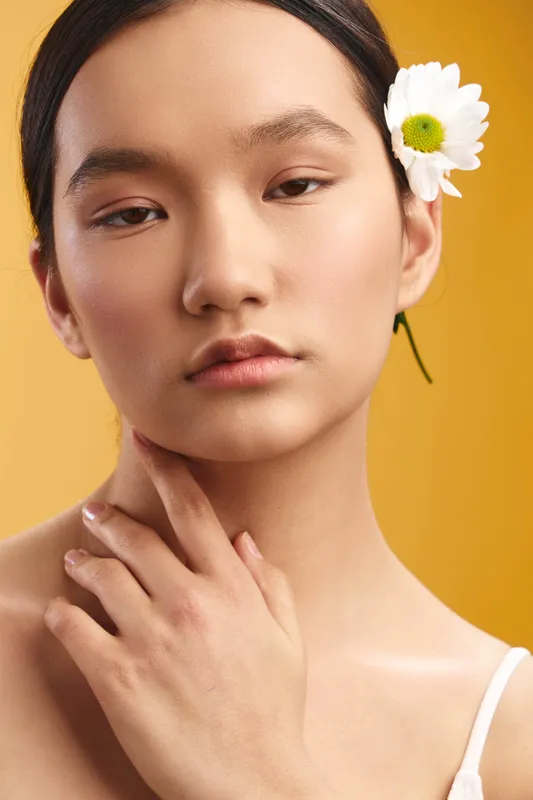 Asian woman with a delicate flower tucked behind her ear poses softly in a studio, radiating calm poise and natural beauty against a warm yellow backdrop.