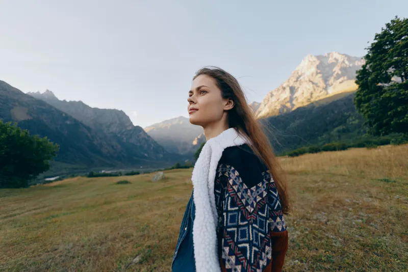 Woman portrait in mountains meadow, nature outdoors scene with young female in patterned shearling coat, looking in profile at scenic landscape under soft sunlight and peaceful atmosphere