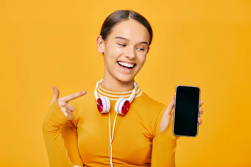 Woman in athletic orange top with headphones, holding a smartphone, smiling against a bright orange background, showcasing fitness lifestyle, sport motivation and digital connectivity for wellness