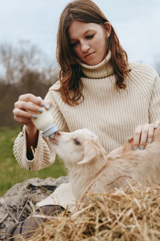 A young woman in a cream knit sweater feeds a newborn goat with a bottle in an outdoor setting. A warm, candid lifestyle moment highlighting animal care and rural living.