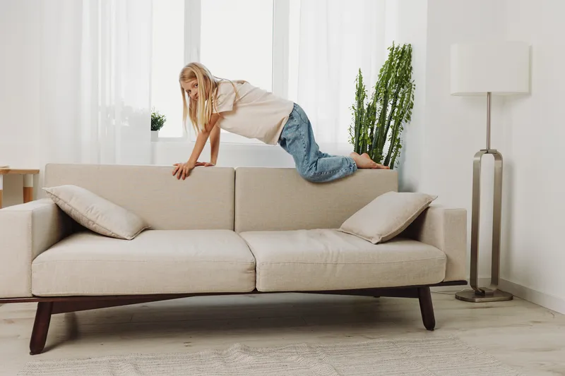 Blond girl climbing on beige sofa in bright living room with plants and white curtains. Concept of childhood play, home activity, and carefree moments indoors.