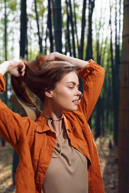 Woman portrait in forest during autumn wearing an orange jacket, hands in hair and eyes closed, natural light and serene expression, outdoor lifestyle and seasonal fashion.
