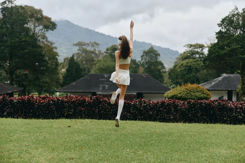 Young woman in a white outfit joyfully dancing in a green field, surrounded by lush nature and mountains, perfect for seasonal and outdoor lifestyle concepts