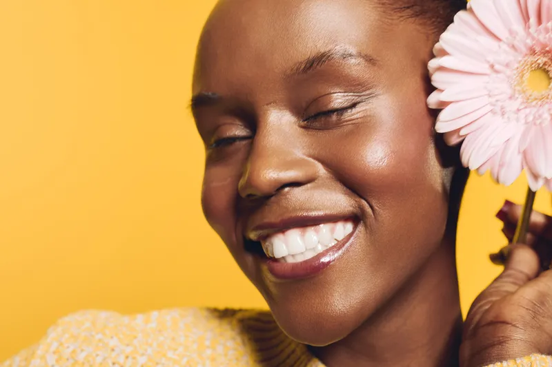 Close-up of happy smiling African woman with dark skin holding pink flower near face on yellow background. Concept of joy, beauty, natural skincare, and wellness with vibrant warm colors.
