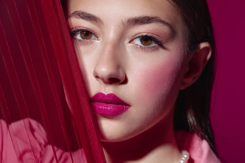 Close-up of a fashion model with bright fuchsia lipstick peeking from behind red fabric on a dark magenta background. Concept of beauty creative and fashion editorial with subtle makeup.