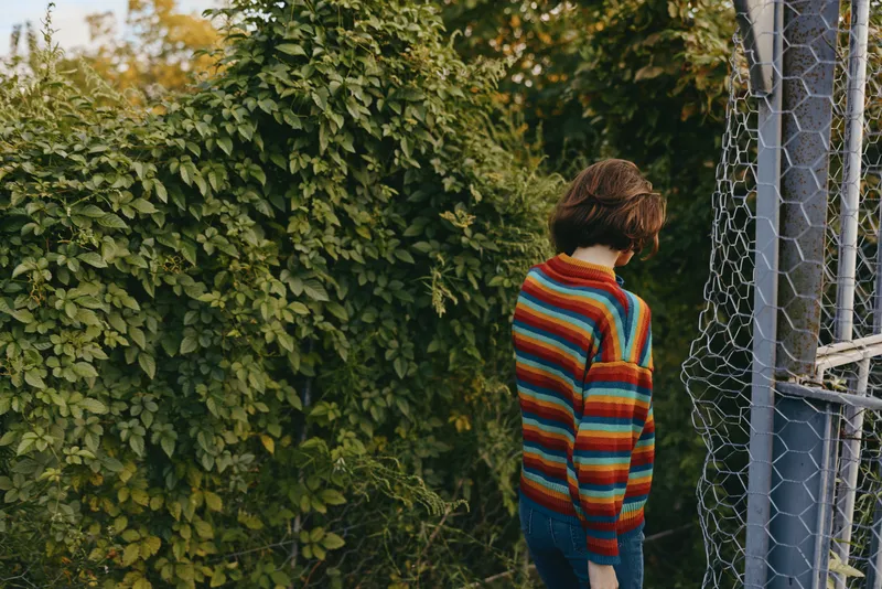 Striped sweater young woman stands contemplative by a chain link fence and overgrown greenery, casual outfit and jeans, short hair, looking down in a moody outdoor lifestyle portrait.