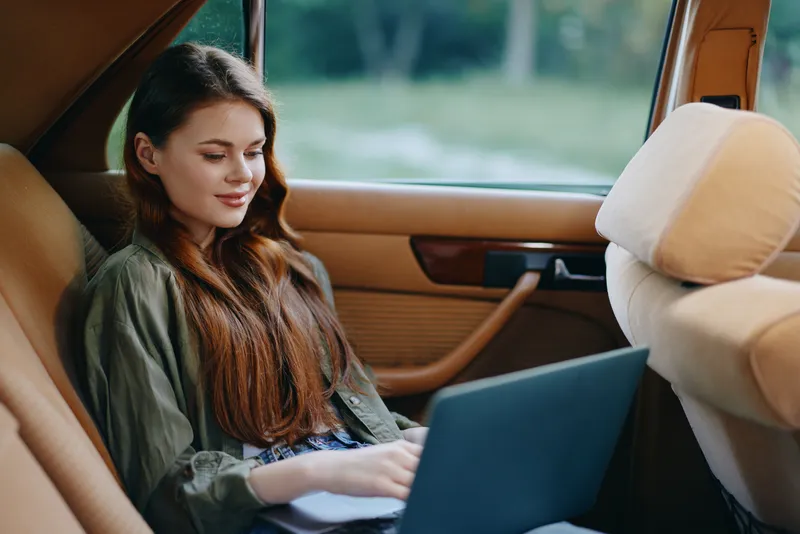Young woman working on a laptop in a vintage car, smiling casually while enjoying the serene outdoor background. Perfect for remote work and lifestyle themes.