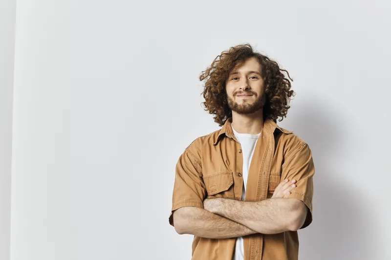 Smiling young man with curly hair wearing a casual brown shirt, standing confidently against a minimalist background, ideal for lifestyle and personal branding visuals
