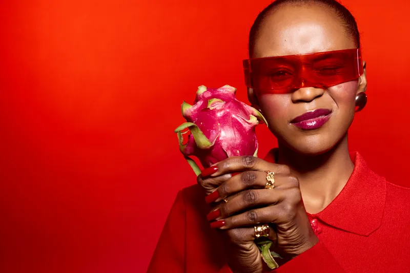 Confident African woman with dark skin wearing red sunglasses and sweater holds a dragon fruit against a vibrant red background. Concept of fashion, fruit, and bold style.