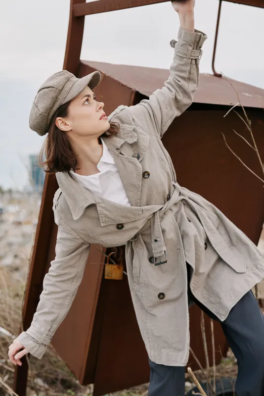 Young woman in beige trench coat and hat climbs rusty metal structure outdoors. Concept of urban exploration, fashion, and autumn casual style with natural background.