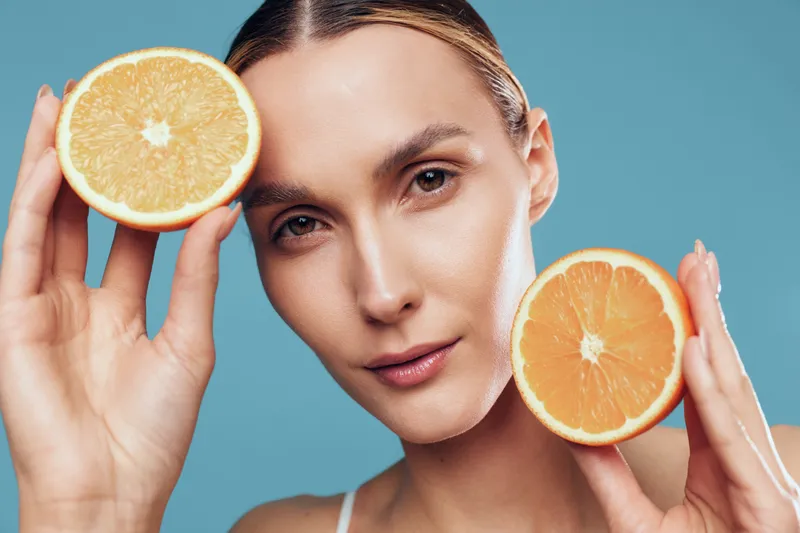 A woman holds orange halves near her face in a bright studio, highlighting fresh citrus and radiant skin. This clean, vibrant image conveys health, beauty, and natural glow.