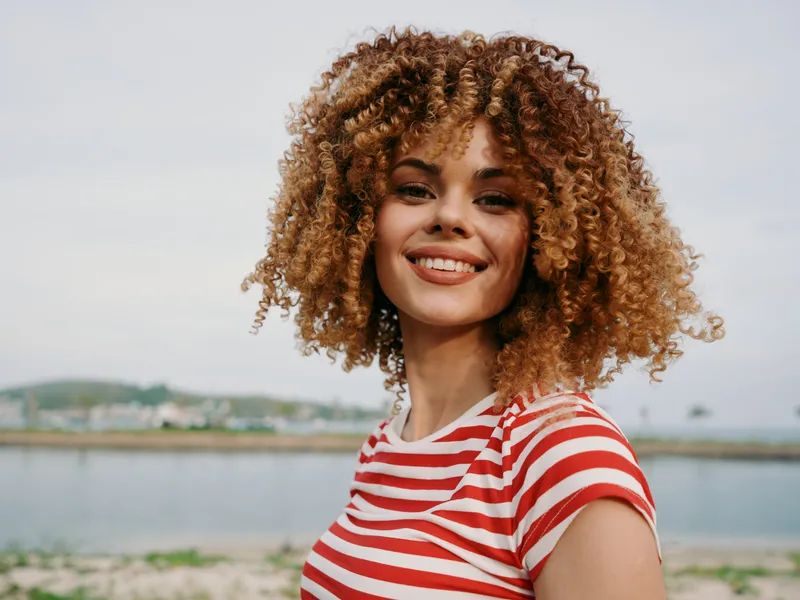 Bright portrait of a smiling woman with curly hair and striped red and white tee, standing outdoors by a calm waterfront, sunny day, natural confidence in casual style perfect for lifestyle imagery.