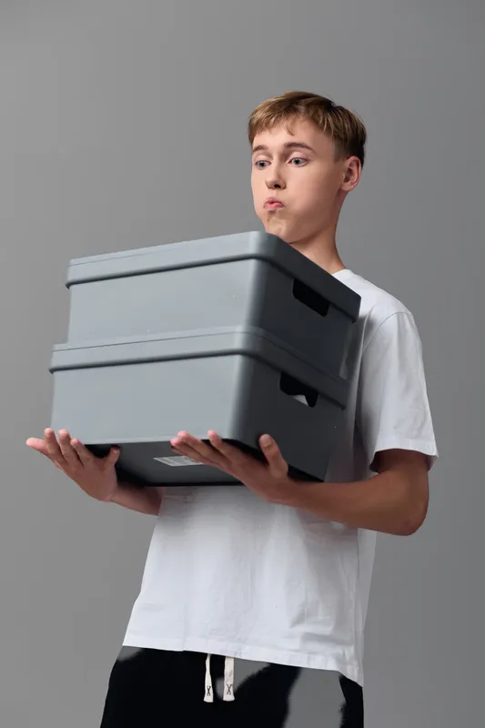 Young man holding a stack of gray storage boxes in a studio, casual white shirt, conveying organization, transport, and delivery readiness with a simple neutral background