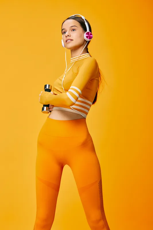 Athletic woman in vibrant orange fitnesswear holds dumbbells, headphones on, posing against a bold orange backdrop for a high-energy workout and training session.
