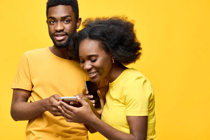 Joyful young African couple sharing fun moments while using a smartphone, dressed in matching yellow outfits against a vibrant yellow background
