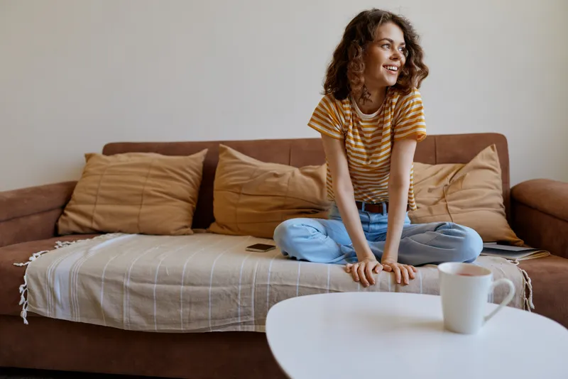 Young woman with curly hair sits cross legged on a cozy living room couch, wearing a striped t shirt and jeans, smiling softly in a warm home moment.