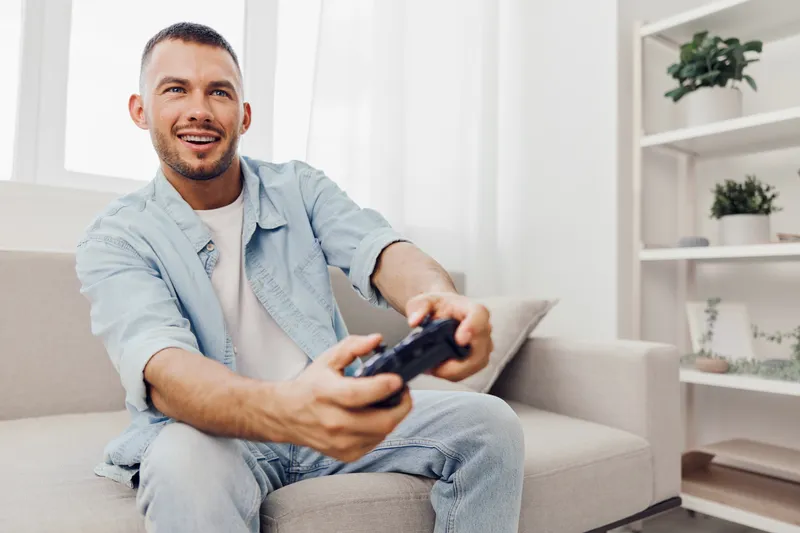 Happy man playing video games in a bright living room, showcasing a modern lifestyle and the joy of gaming. Ideal for tech and leisure themes in 2025-2026.