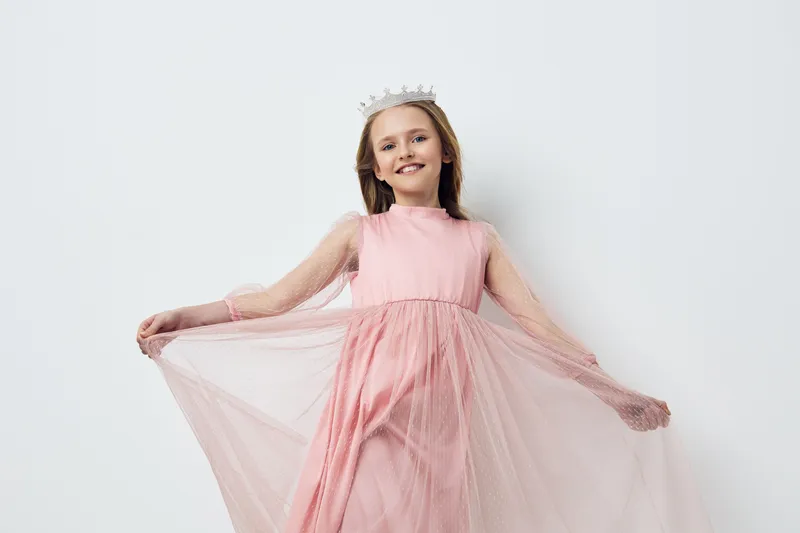 Smiling young girl in a pink dress with tulle sleeves and a crown, posing joyfully against a neutral background, ideal for childhood and fashion concepts