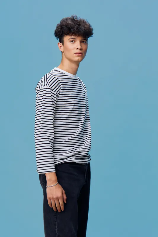 young man wearing striped long sleeve shirt and black jeans standing against blue background with serious expression and curly hair, casual style portrait