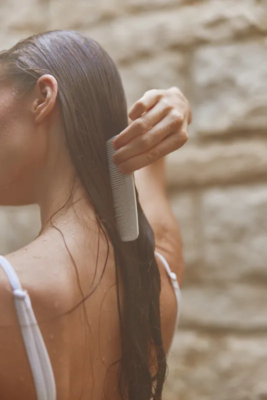 Woman combing wet long hair with a white comb, showing smooth texture and natural shine in soft natural light with blurred beige stone background.