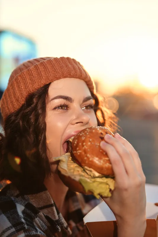 Woman eating a burger at sunset in beanie, candid outdoor portrait capturing authenticity and golden hour glow, mindful living vibe with emotional storytelling and genuine enjoyment.