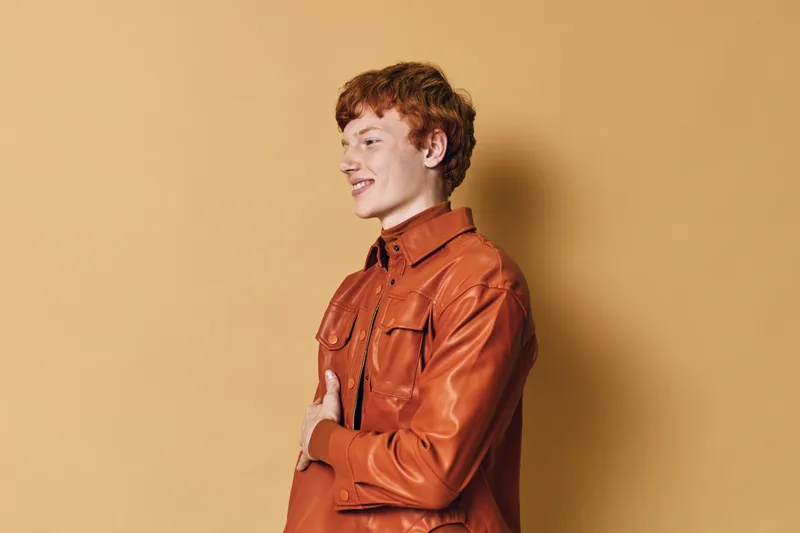 Young man with red hair and freckles wearing brown leather jacket standing sideways and smiling on beige background. Casual style, fashion portrait, confident and happy male model.