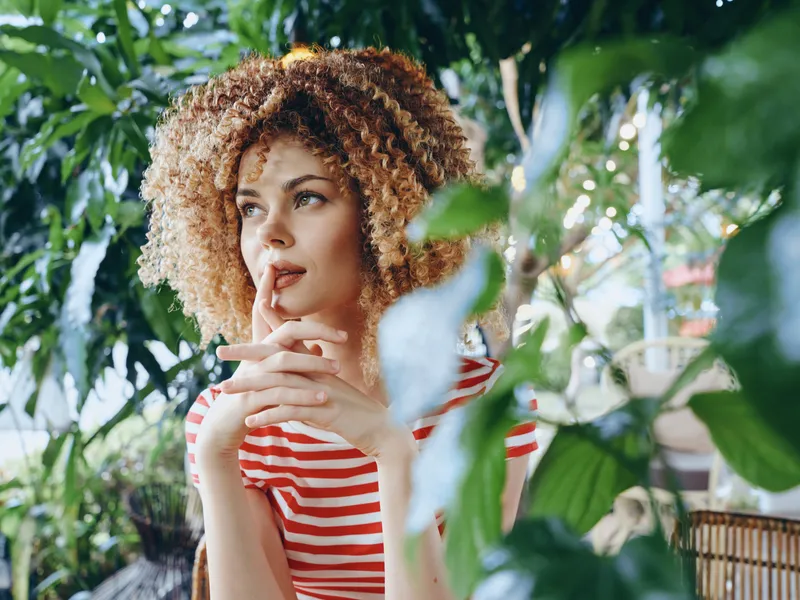 Thoughtful woman with curly hair stands among lush green leaves, wearing a red striped shirt, fingers near lips, gazing aside as sunlight filters through foliage.