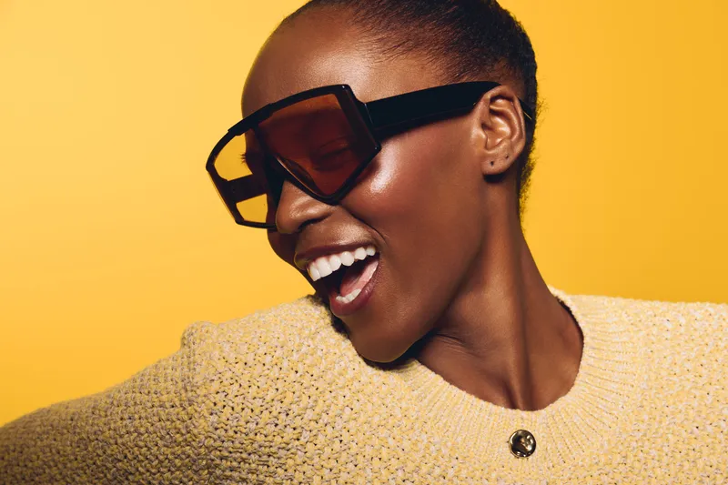 Stylish African woman with dark skin wearing oversized sunglasses and beige sweater smiles joyfully on yellow background. Concept for fashion, beauty, and positive mood portrait.
