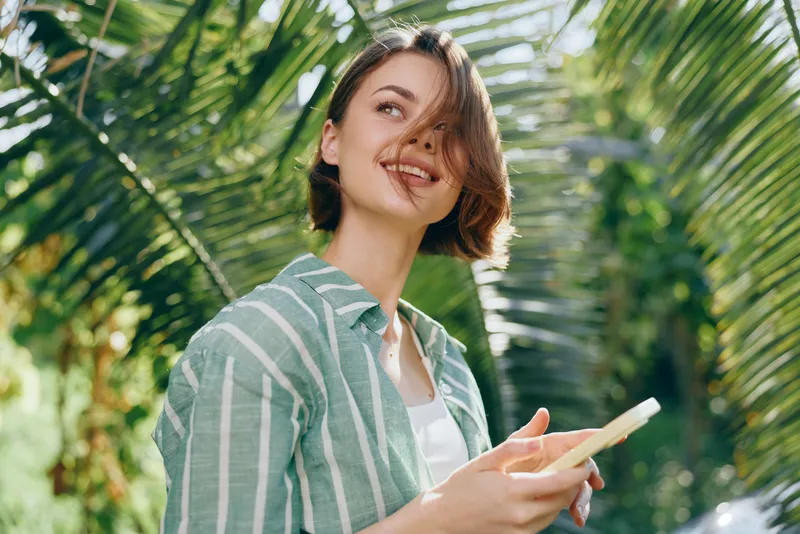 Smiling woman in a striped shirt stands outdoors among lush greenery, holding a device, under bright natural light, a warm candid portrait.