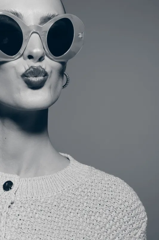 Stylish young caucasian woman with pale skin wearing large sunglasses and textured sweater blows a kiss on grey background. Fashion portrait with retro glamour and confident mood.