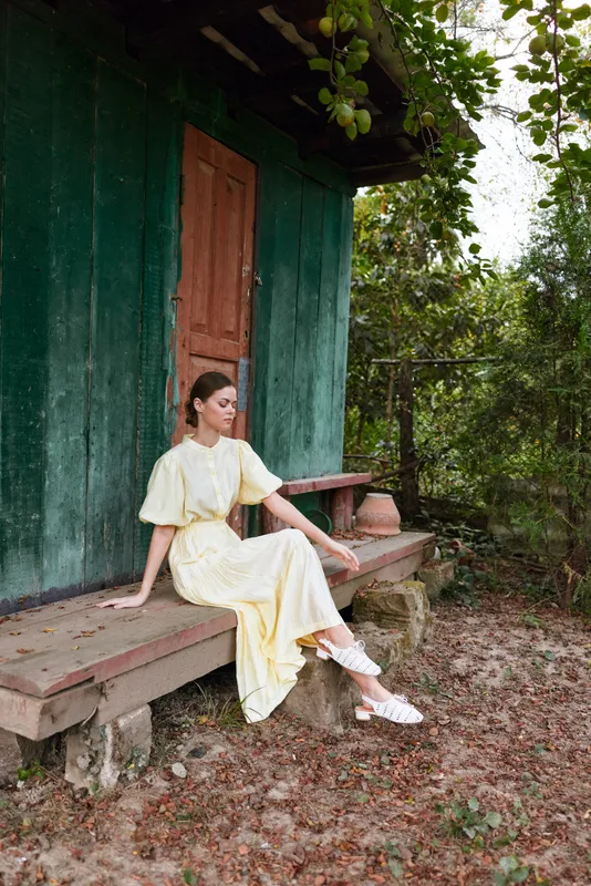 A woman in a pale yellow dress sits on a wooden bench outside a green rustic cabin, surrounded by trees and gravel, presenting a calm countryside moment.