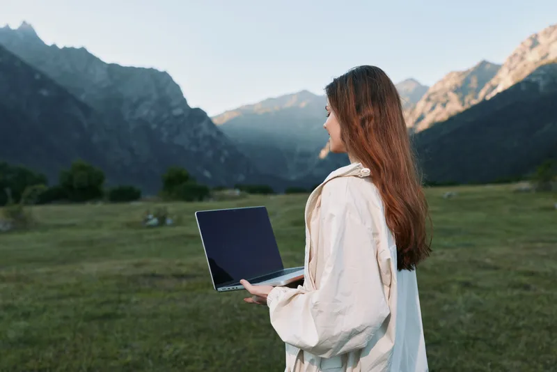 A woman stands outdoors with a laptop in a scenic valley and mountains in the background. She faces sideways, exuding focus and calm while enjoying a productive moment.