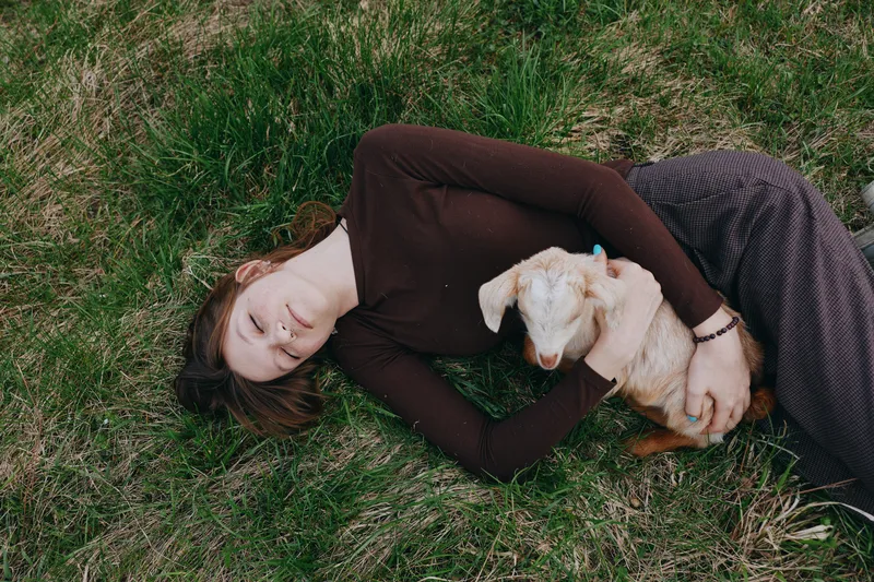 Young woman with long brown hair lies on the grass gently holding a small white baby goat, enjoying a peaceful moment in a natural outdoor setting with soft daylight.