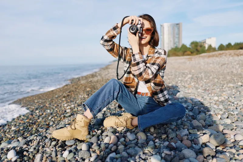 Woman photographer on a rocky beach with a camera, capturing seascape and sky, wearing casual plaid shirt, jeans and boots, enjoying outdoor shoot in daylight.