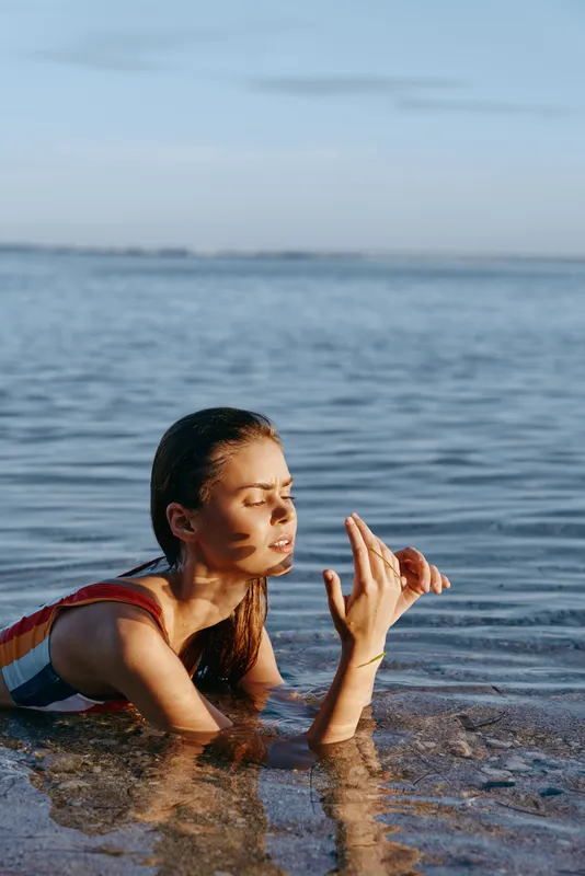 Woman ocean beach swimwear sunrise nature wellness freedom relaxation, kneeling in shallow water with a relaxed smile, hands raised in a calm gesture, soft blue horizon, peaceful seaside
