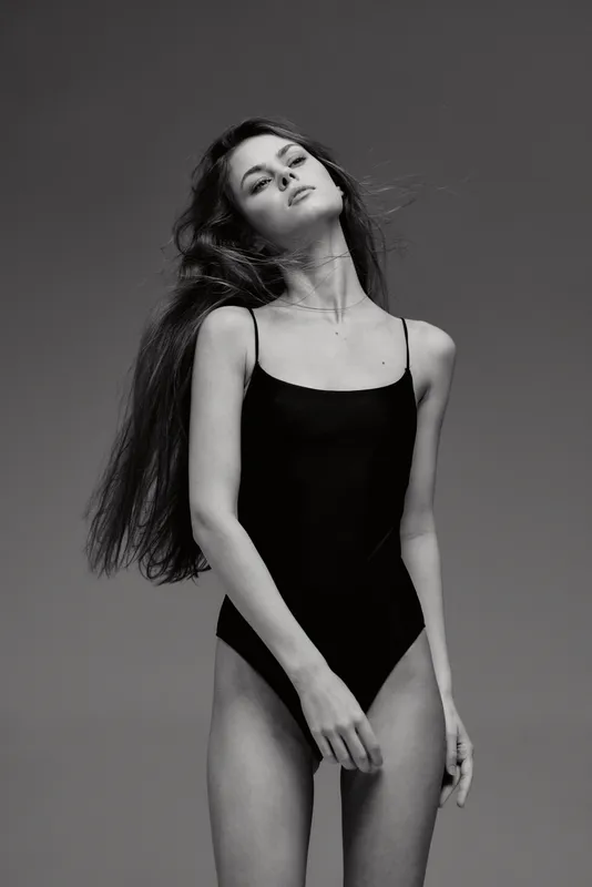 Black and white portrait of a slender woman in a black bodysuit with long hair posing gracefully in studio lighting.