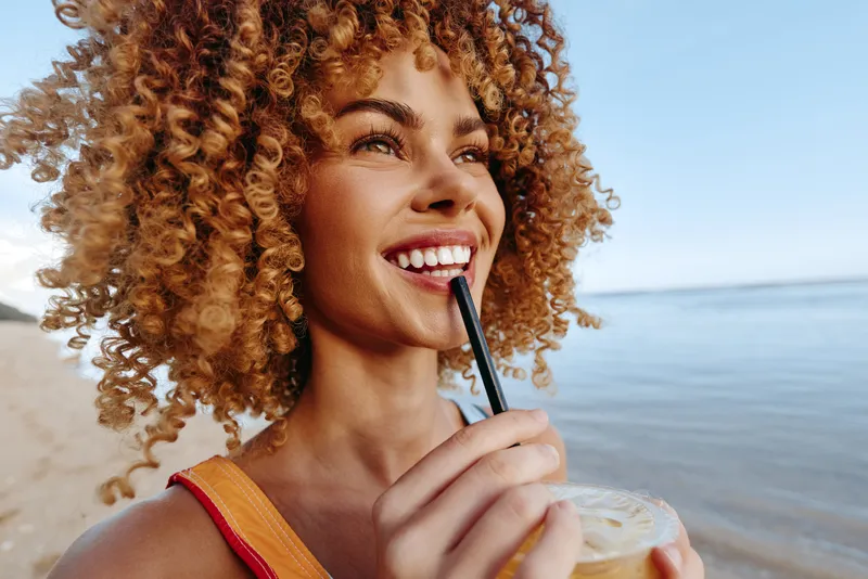 Young woman with curly hair enjoys a drink with a straw on the sunny beach, smiling as she looks toward the ocean.