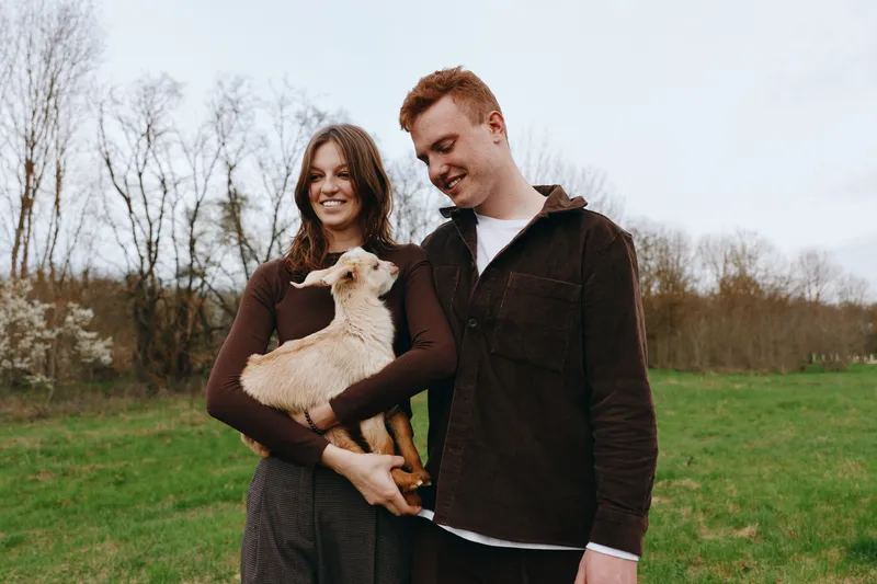 Casual couple outdoors with a light brown dog in a green field, they share warm smiles and affection, showcasing a relaxed lifestyle in a natural countryside setting