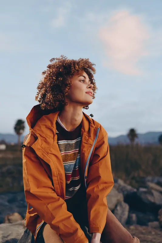 Woman lifestyle portrait outdoors, curly hair, orange jacket, sky, mountains background, calm relaxed mood, woman looking upward, standing on scenic landscape in fresh daylight