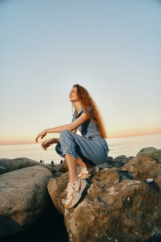 young woman with long curly hair sitting on rocks near the sea during sunset in casual clothes and sneakers, enjoying peaceful outdoor moment
